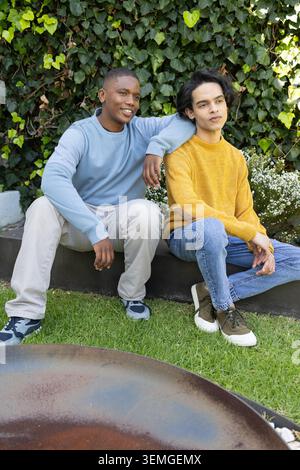 Diverse male friends sitting on low planter in garden in sweaters near rusted fire pit Stock Photo