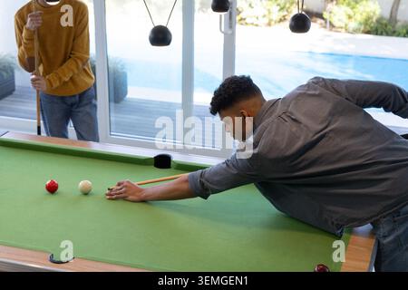 Two men leaning over pool table in rec room, aligning cue toward sliding door and pool Stock Photo