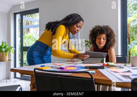 African American women collaborating, mustard, maroon tops using tablet, notes, pens in home studio Stock Photo
