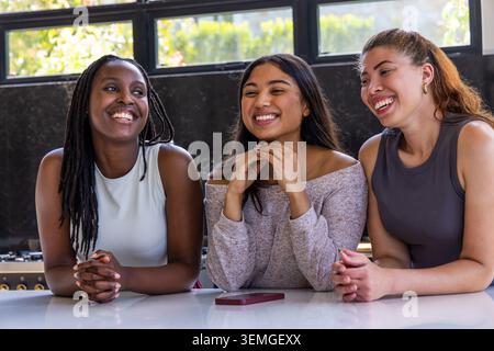 Diverse female friends laughing and leaning forward at kitchen counter with maroon smartphone Stock Photo