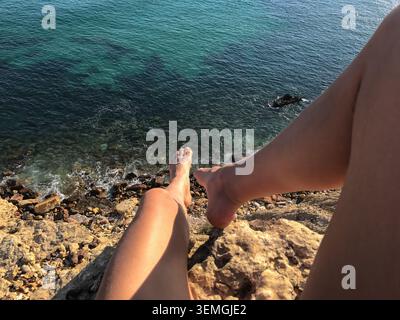 Low-angle perspective of an unrecognizable woman sitting on golden sandstone cliffs high above the Atlantic Ocean. Her legs extend toward camera. Stock Photo