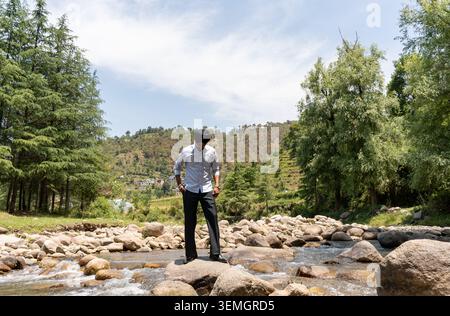 A wide horizontal shot of a young Sikh man in formal clothing looking down thoughtfully while posing on rocks in a rushing mountain river. Stock Photo