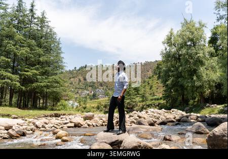 A young handsome Sikh man wearing a traditional turban and formal clothing stands confidently on rocks in a rushing river. Stock Photo