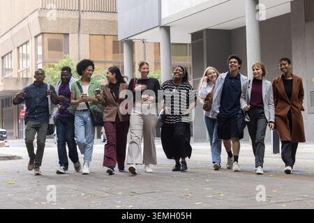 Diverse friends walking together on wide city sidewalk, carrying coffee cups and backpacks Stock Photo