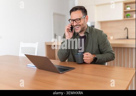 Smiling man connecting remotely. Laughing during a phone call. Holding a coffee mug. And working on a laptop at a bright home office table. Embodying Stock Photo