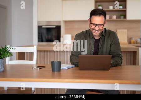Smiling man finding job satisfaction while telecommuting, operating a laptop in his modern kitchen, embodying concepts of flexibility, productivity, a Stock Photo
