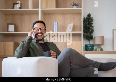 Man wearing eyeglasses comfortably relaxing on an armchair, engaging in a happy phone conversation, enjoying a hot beverage, and embracing a modern ho Stock Photo