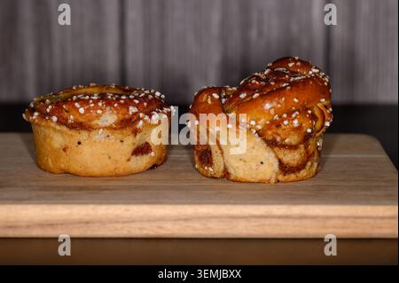 Two cinnamon rolls sit on a wooden board in a kitchen. One roll features pastry cream on top, while the other has a traditional swirl. The setting is Stock Photo