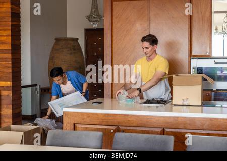 Diverse male friends unpacking framed artwork, boxes, bubble wrap and smartphone on kitchen island Stock Photo