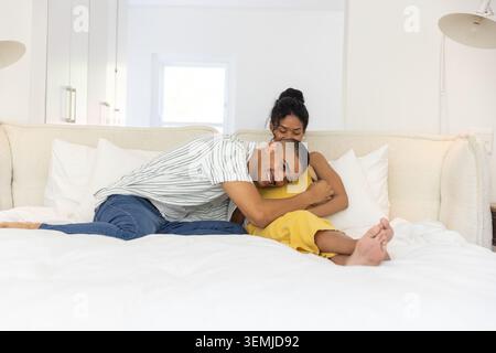 Couple cuddling on white bed in bedroom, woman in yellow dress, man wearing striped shirt, jeans Stock Photo