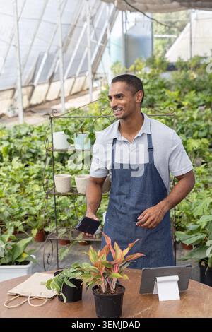 Indian man wearing grey polo, dark apron, using phone, tending plants on wood table in nursery Stock Photo