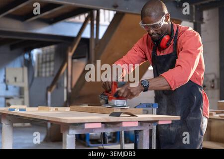 African American man operating sander smoothing wooden board in workshop wearing apron, copy space Stock Photo