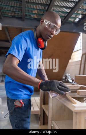 African American male carpenter standing at bench wearing goggles smoothing wood with sanding block Stock Photo