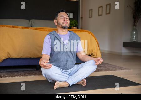 Mature Asian man sitting meditating on black mat in bedroom near mustard duvet wearing grey hoodie Stock Photo