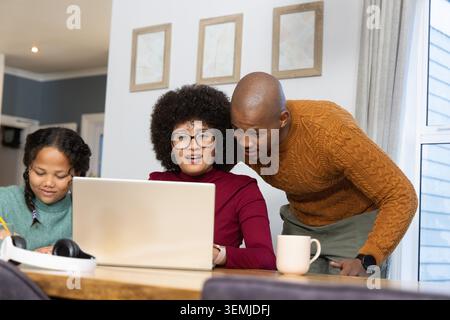Diverse family parents and daughter sitting at dining table, woman working on laptop, child writing Stock Photo