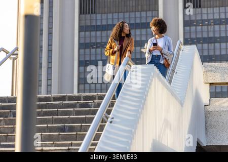 Diverse female friends walking down concrete stairs at urban plaza, holding smartphone and bags Stock Photo