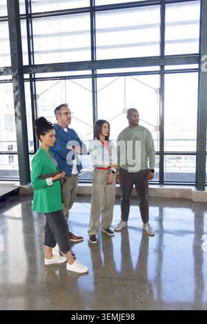 Diverse coworkers standing and touring office by large windows with tablet, floor reflections Stock Photo