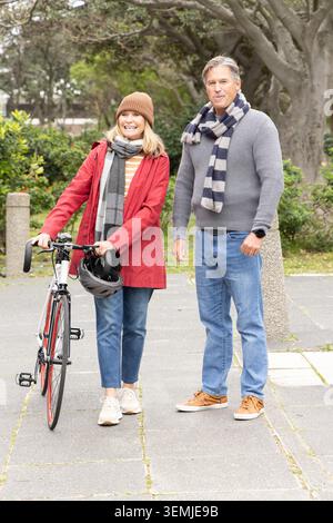 Senior couple standing in park on walkway, woman wearing red coat holding road bicycle and helmet Stock Photo
