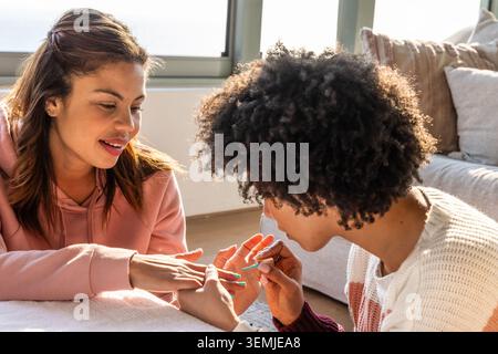 Diverse female friends sitting by window at home, painting fingernails with polish brush Stock Photo