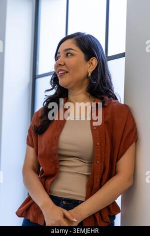 Woman leaning against white wall in studio by tall window, wearing rust shirt and gold hoops Stock Photo
