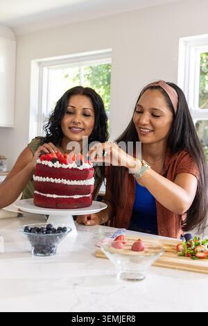 Diverse female friends on marble island decorating red velvet cake on cake stand with strawberries Stock Photo