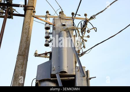 High voltage pole mounted transformer with multiple electrical wires stands against a clear sky. Substation distribution equipment provides grid power Stock Photo