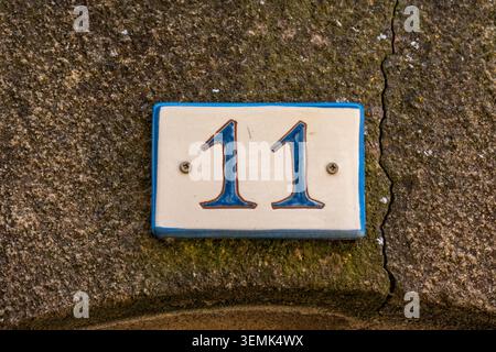 A blue and white ceramic house number 11 plaque screwed onto a cracked and mossy weathered stone wall in a French village Stock Photo