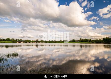 A picturesque lake perfectly reflects a bright blue sky filled with massive, puffy clouds. On the distant shore, small buildings and a line of trees c Stock Photo