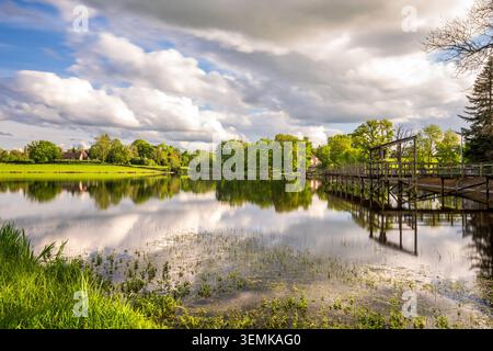 A long wooden walkway and pier wind along the shore of Les Pouyades lake, reflecting in the calm water. The dense line of green trees and large white Stock Photo