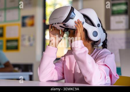 School-aged child putting on white VR headset at desk in classroom wearing light pink sweatshirt Stock Photo