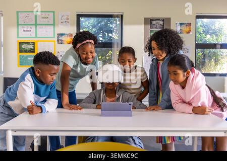 Diverse children gathering around seated student wearing white VR headset, watching tablet in class Stock Photo