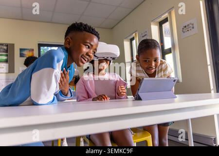 Three African American children gathering around white table at school using VR headset and tablet. Classroom, teamwork, technology, learning, educati Stock Photo