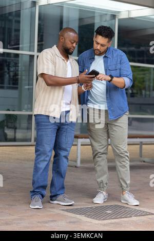 Two African American men standing on paved plaza outside modern glass building checking smartphone Stock Photo