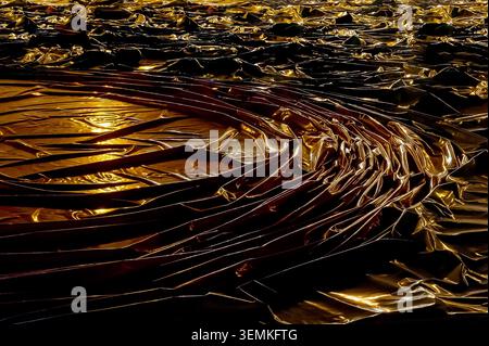 Dark, reflective material with numerous folds and wrinkles, dramatically highlighted by a warm, yellow light source Stock Photo