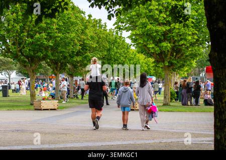 A family with a child on shoulders walks along a tree-lined path at a busy outdoor food and drink festival in Southport, UK. Stock Photo