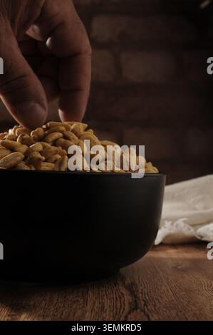Male hand fingers taking roasted peanuts from a black bowl, with a white cloth napkin and brick wall in the background, vertical baroque chiaroscuro Stock Photo