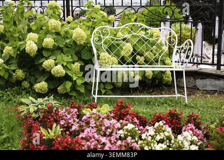 White metal bench with hydrangea flowers in a lush garden with a black wrought iron fence creating a quiet and picturesque scene Stock Photo