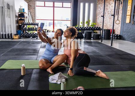African American female friends in athletic wear taking selfie on yoga mats in studio holding phone Stock Photo