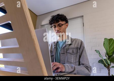 Asian man standing at slatted home workstation wearing denim jacket, eyeglasses, working on laptop Stock Photo