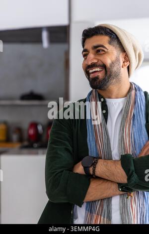 Adult man standing arms crossed, smiling looking right in kitchen wearing beanie, striped scarf Stock Photo
