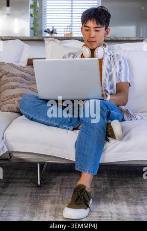 Asian male teenager studying on white sofa at home with silver laptop and white headphones. Solo, student, focus, cozy, minimalist, daylight, kitchen Stock Photo