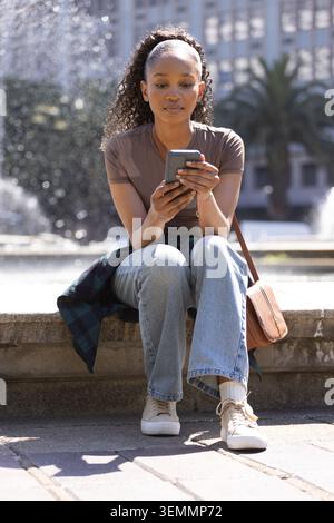 African American woman in her 20s sitting on stone fountain edge holding smartphone, wearing jeans Stock Photo