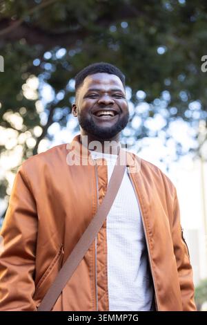 Adult man standing smiling in park, wearing rust bomber jacket, white textured sweater, tan strap Stock Photo
