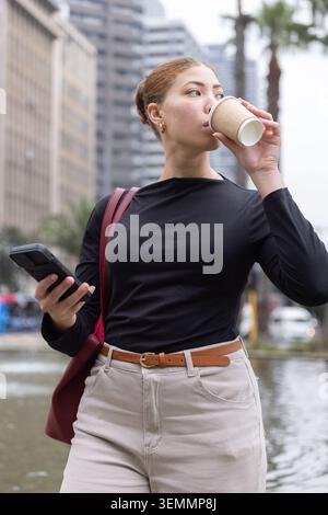 African American woman in black top and pants drinking from paper cup checking phone by fountain Stock Photo
