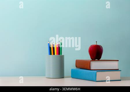 A pencil holder filled with brightly colored pencils next to a red apple on a stack of two books. Education, back-to-school season, creativity Stock Photo