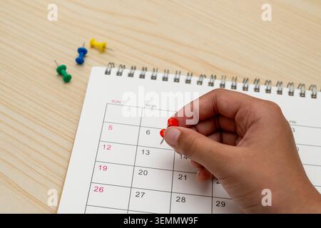 A  hand pressing a red pushpin into a specific date on a calendar. Scheduling, deadlines, and time management concept. Copy space for text. Stock Photo