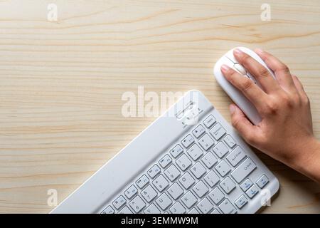 A top-down flat-lay view of a hand operating a white wireless computer mouse next to a white keyboard on a light wooden desk. Stock Photo