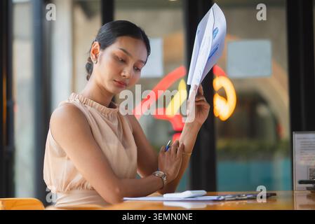 Focused businesswoman reviewing printed report with pen at office cafe desk, analyzing charts and documents for accounting, budgeting, market research Stock Photo