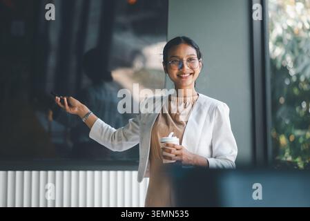 Smiling businesswoman holding coffee cup and remote while presenting in office meeting, combining business communication, confident leadership and rel Stock Photo
