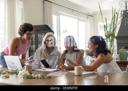 Intergenerational female group reviewing documents, using phone, laptop, tablet at dining table Stock Photo